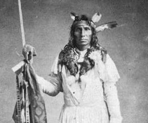 Little Crow in three quarter height view wearing a headress with three feathers and carrying a spear [5]