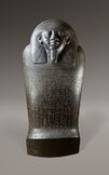 Black and white image of a dark stone coffin with a human face, the coffin stands upright facing the viewer.