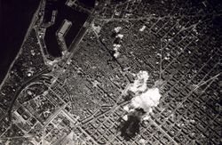 Black and white photo of the city from high above. Smoke from a bomb can be seen