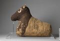 Cattle mummy representing a seated animal. The head is lifelike, while the body is rendered as a rough trapezoid. The head and forepart of the body are covered in a dark brown fabric, while the rest of the animal is covered with an undyed cloth. Underneath the undyed cloth, several other textile fabrics have been used as a stuffing.[35]
