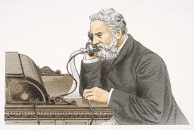 A grizzly and well-dressed Alexander Graham Bell sits at a desk talking over a candle-stick telephone, surrounded by numerous business executives and news reporters who are witnessing a historic event in the atrium of a large corporate building.
