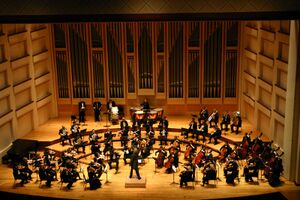 Orchestra, in evening dress, performing on a pine-panelled stage