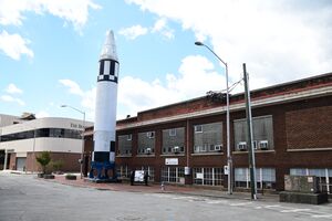 An exterior photo of an upright rocket standing outside of a brick building