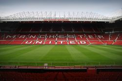 A stand of a football stadium. The seats are red, and the words "Manchester United" are written in white seats. The roof of the stand is supported by a cantilever structure. On the lip of the roof, it reads "Old Trafford Manchester".