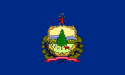 The flag shows a gold-colored heart-shaped frame around a scenic illustration. In the center there is a tall spruce-like tree, surrounded by grassy fields with a wandering cow and hay bales. In the background, tall mountains are visible. Above this frame and illustration is a deer head and surrounding it is a wreath of leaves. Below it, text reads "Freedom, Vermont, and Unity" in white.