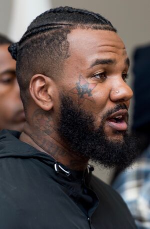 A black man with face tattoos and cornrows, speaking formally at an off-screen entity
