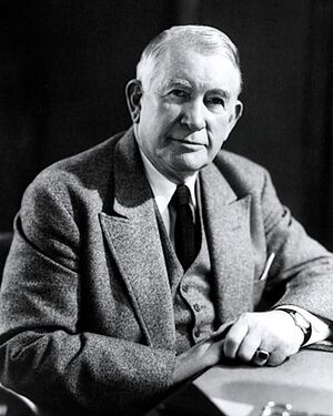 A man with white hair wearing a gray jacket and vest, black tie, and white shirt, seated and leaning on a desk