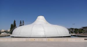 A gray semi-hemispherical structure with a protruding top, standing in the middle of a fountain, with several jets of water falling on the structure