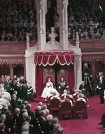 Queen Elizabeth II and Prince Phillip sit on thrones before a full Parliament.jpg