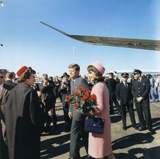 Kennedy and the First Lady, dressing in a pink outfit and holding a bouquet of flowers, depart from Air Force One and greet welcomers
