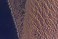 The dune sea of the Namib Desert. The crests of the dunes are aligned in a marked northwest-southeast orientation. The dunes act as obstacles, causing the winds to be deflected significantly to the right, in effect reorienting the southerly wind to become a southwesterly wind. A strand plain lies between the dunes and the ocean.