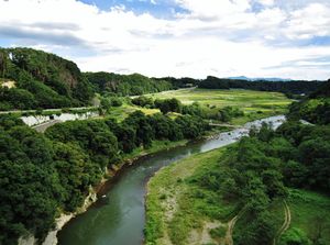 River moving left to right in the mid ground surrounded by green grass and trees and mountains in the background.