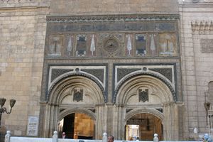 Two large arches recessed into a wall are visible, with entrance-ways at their bottom. Above the arches the wall is ornately decorated with carvings and geometric designs. Parts of the decorations are colored red, blue and gold. The arches and decorations are surmounted and flanked by large, closely-set rectangular blocks.