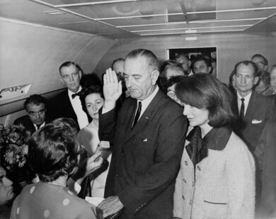 Lyndon B. Johnson raises his hand above an outstretched Bible as he is sworn in as President as Air Force One prepares to depart Love Field in Dallas. Jacqueline Kennedy, still in her blood-spattered clothes (not visible), looks on.