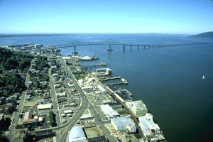 A long bridge crosses a huge river flowing into a vast body of water under a blue sky. The bridge begins in a settlement with streets, buildings, and wharves along the river and extends out of sight toward a low hill on the far shore. The first part of the bridge has a superstructure and is high above the water but then the bridge gradually descends and continues out of sight not so far above the water.