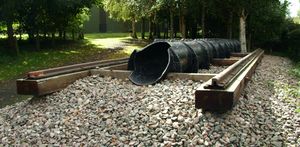 Exterior in woodland. a short section of railway line on wooden sleepers with a cast iron pipe of approximately one foot diameter, running inline with the rails